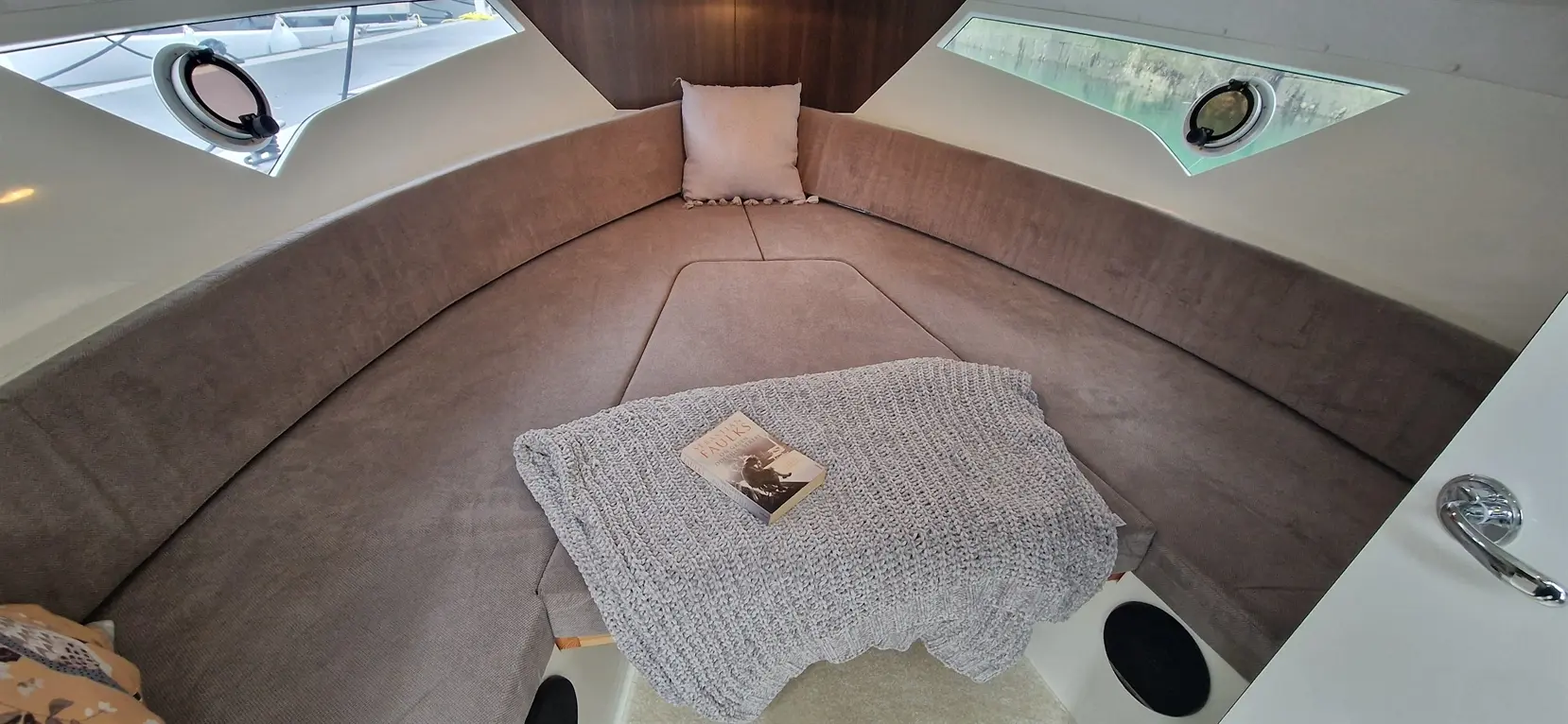 A cozy boat interior features plush grey seating, a book on a grey blanket, and oval windows revealing water views.